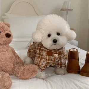 Chien blanc bouclé portant un manteau à carreaux bruns avec bouton effet bois, posé sur un lit