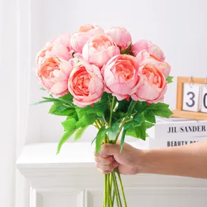Mano sosteniendo un frondoso ramo de peonías rosas artificiales con hojas verdes en un interior luminoso