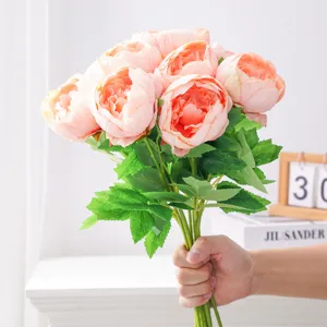 Ramo de peonías rosa anaranjado sostenido en la mano, con hojas y tallos verdes sobre fondo claro