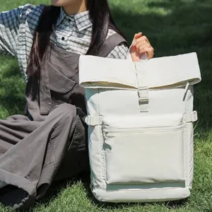 Ampio zaino avorio con chiusura roll-top e tasca frontale con zip, mostrato all’aperto sull’erba