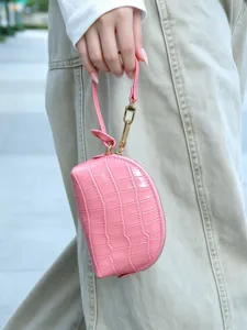 Close-up of a pink embossed croc-texture wrist pouch with gold hardware held beside beige trousers