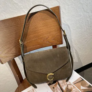 Borsa in suede verde oliva appesa a una sedia di legno, con tracolla regolabile e chiusura dorata