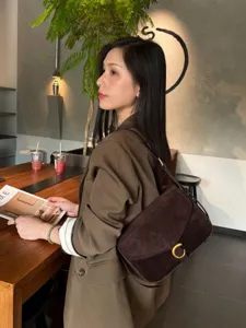 Borsa a spalla marrone scuro effetto suede, con anello dorato, indossata accanto a un tavolo