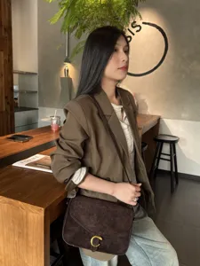 Borsa in suede marrone scuro con chiusura dorata a C, indossata a tracolla da una modella seduta