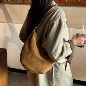 Sac bandoulière demi-lune marron effet suédé, à sangle tressée, porté sur un trench clair