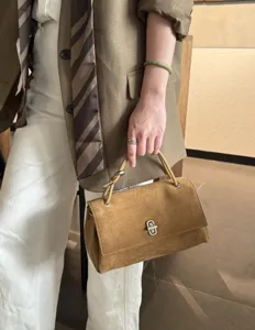 Close-up of a hand holding a tan suede handbag with a matching handle and silver turn-lock clasp