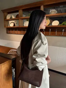 Woman carrying a dark brown suede shoulder bag beside a wooden counter in warm indoor light
