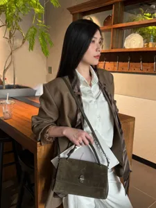Model carrying a dark brown suede flap bag with a metal turn-lock in a warm café setting