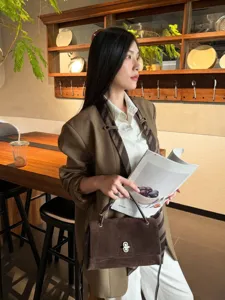 Lifestyle view of a deep brown suede bag with a metal clasp, carried by a seated woman in a café