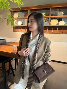 Lifestyle view of a dark brown suede flap bag with metal clasp, worn crossbody in a café