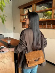 Bolso marrón efecto ante con acolchado de rombos y cadena plateada, llevado al hombro en un café