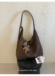 Sac tressé marron avec pendentif ourson, présenté sur un tabouret noir devant un mur blanc