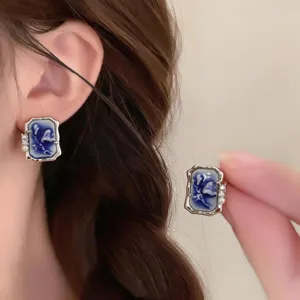 Close-up showing a blue-white square earring worn on the ear and held by hand, with pearl accents