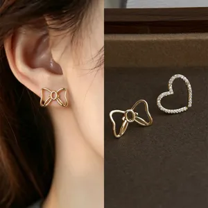 Close-up of a gold bow stud worn on an ear, beside a gold open-heart stud with clear stones
