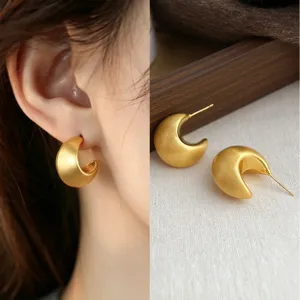Close-up of a matte brushed gold crescent earring worn on the ear, with its matching pair displayed flat