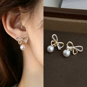 Gold bow earrings with clear stones and dangling white pearls, worn and displayed on velvet.