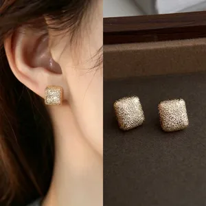 Close-up of a textured gold-tone square stud on an ear, with its pair on a brown velvet tray