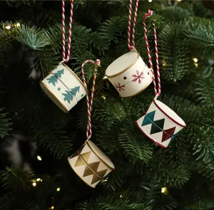 Four cream metal drum ornaments with festive patterns hang among evergreen branches and warm lights.