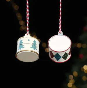 Two painted metal drum ornaments with tree and diamond patterns hang against warm festive lights