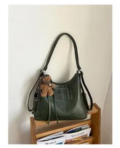 Deep green faux-leather bag with stitched panels, a long strap and a small teddy bear charm