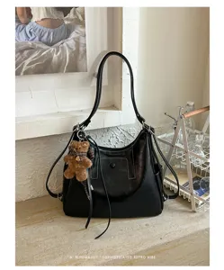 Black leather-look bag with stitched panels and a brown teddy bear charm, displayed by a window.