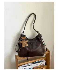 Dark brown leather-look bag with stitched flap and plush bear charm wearing a checked bow
