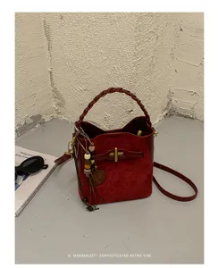 Burgundy suede bucket bag with braided handle, gold clasp and bead tassel, styled on a tabletop