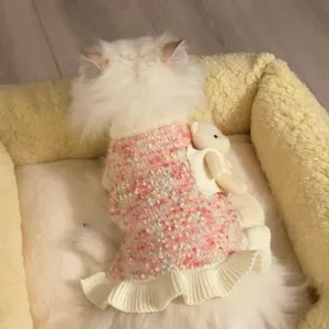 Un gato lleva un vestido de punto rosa jaspeado, con volante acanalado crema y colgante de osito
