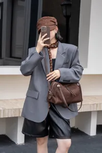 Brown suede-textured handbag with silver hardware styled crossbody over a gray blazer outdoors
