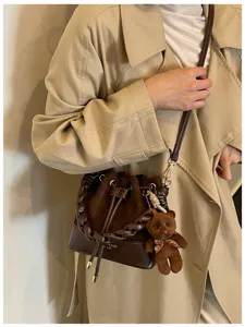 Brown bucket bag with a chunky braided handle and teddy charm, worn over a beige trench coat