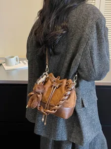 Brown suede-look bucket bag worn crossbody, featuring a braided handle and teddy bear charm
