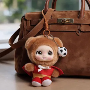 Plush doll keychain with a soccer ball charm hanging from the strap of a brown suede handbag