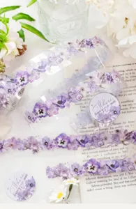 Translucent decorative tape with purple flowers and pearl accents arranged across an open book