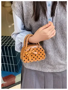 A child holds a glossy tan mini bag with black polka dots and a raised bow, shown on the wrist
