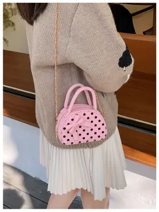 Pink mini bag with black polka dots and a bow, worn on a gold chain over a cream outfit indoors.