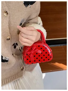 Close-up of a glossy red polka-dot mini bag with a bow, held by hand against a knit cardigan