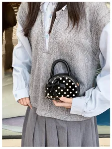 A small glossy black polka-dot bag with a bow, held against a gray vest and blue shirt