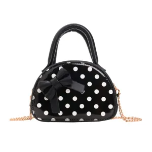 Front view of a glossy black mini bag with white dots, a bow, top handle and gold chain strap