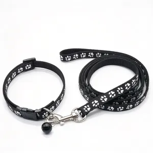 Black nylon dog collar and leash set with silver paw prints, a bell and metal clip, laid out on white