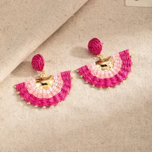 Close-up of pink woven fan-shaped earrings with gold-tone beads displayed on beige linen