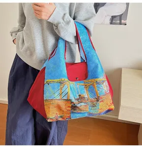 A model carries a blue and red tote with a vivid painterly bridge scene in an indoor setting