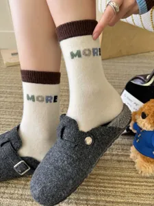Close-up of cream knit socks with brown ribbed cuffs and “MORE” lettering, worn with gray slippers