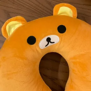 Close-up of an orange plush bear-shaped neck pillow with embroidered features and pale yellow ears