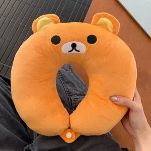 Close-up of an orange plush bear-shaped neck pillow with embroidered face, yellow ears and clasp