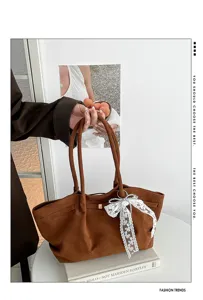Close-up of a brown suede-textured tote with long handles and a white lace bow charm, held indoors
