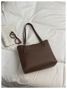 Borsa marrone in similpelle su biancheria da letto bianca, accanto a un libro aperto e occhiali da sole