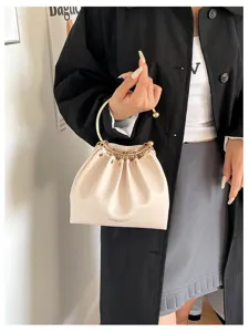 Modella con borsa plissettata color crema, anello dorato e borchie, davanti a una parete chiara.
