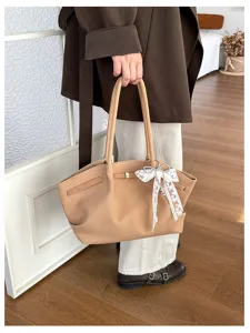 Person holding a camel suede-look tote with gold clasp and white lace bow charm indoors.