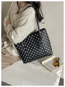 Borsa nera a pois bianchi portata a spalla, accanto a un libro aperto e occhiali da sole