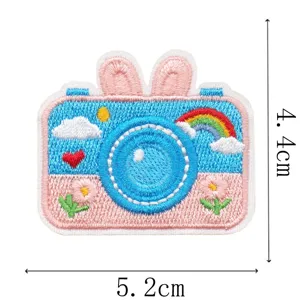 Close-up van een blauw-roze geborduurde cameravorm met konijnenoren, regenboog, wolken en bloemen